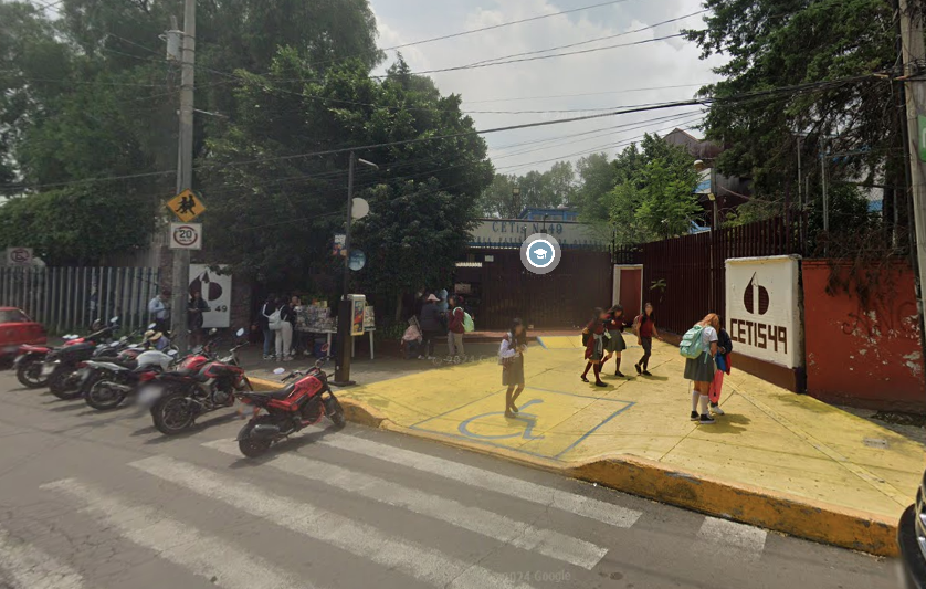 Fotografía en la que se muestra la entrada del plantel, conformada por una explanada a la que se puede ingresar por medio de una rampa. Al fondo de la foto está la puerta de acceso, que es color rojo. A la derecha de la puerta hay un muro de color blanco en el que, dice en letras grandes rojas, CETIS 49.